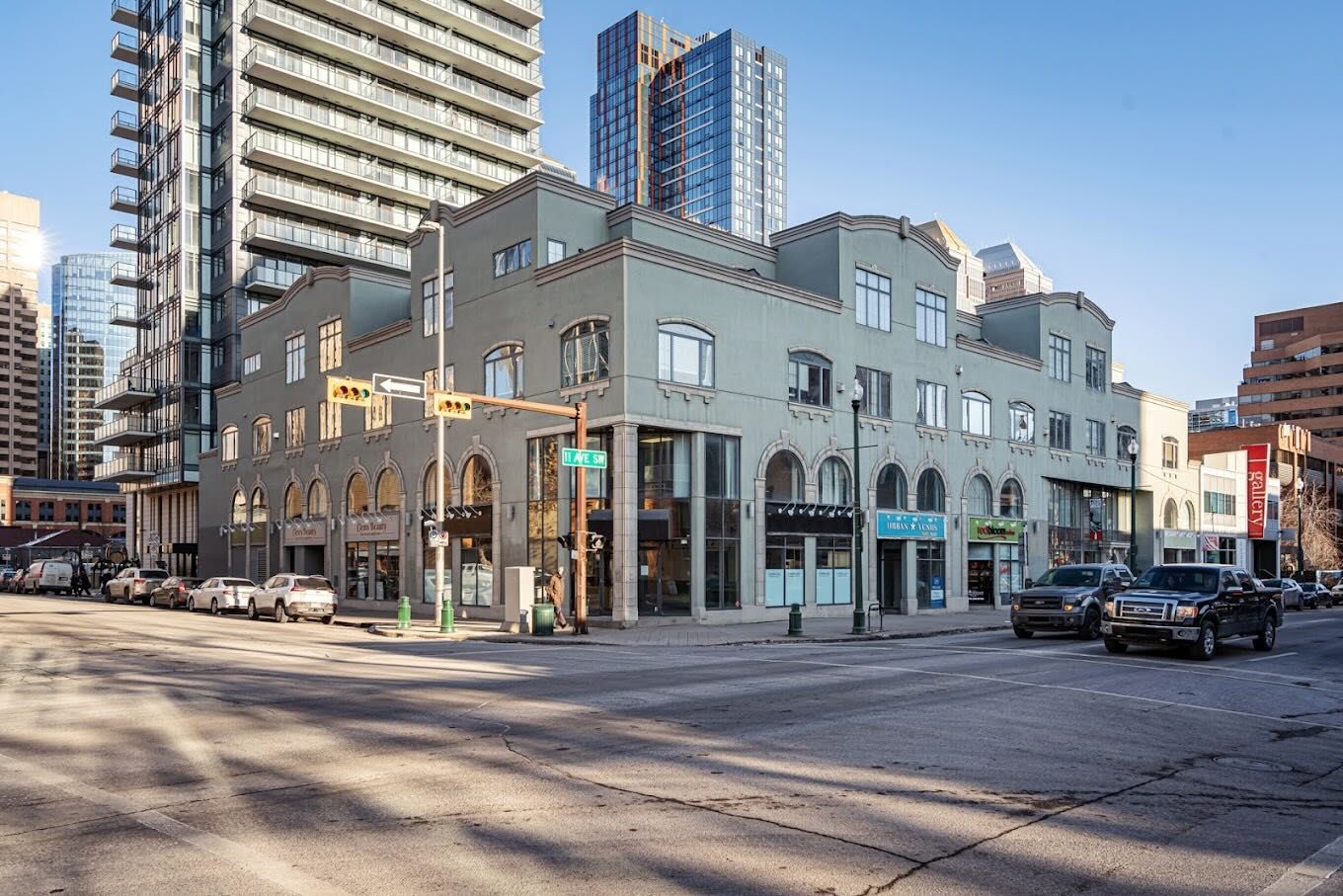 A grey stucco heritage building on the corner of 11 Ave SW in Calgary, featuring arched windows and decorative parapets, with street-level retail shops including Urban Venus. A modern glass condo tower rises directly behind it, and additional high-rises are visible in the background against a clear blue sky. Cars are parked along the street and at the intersection.