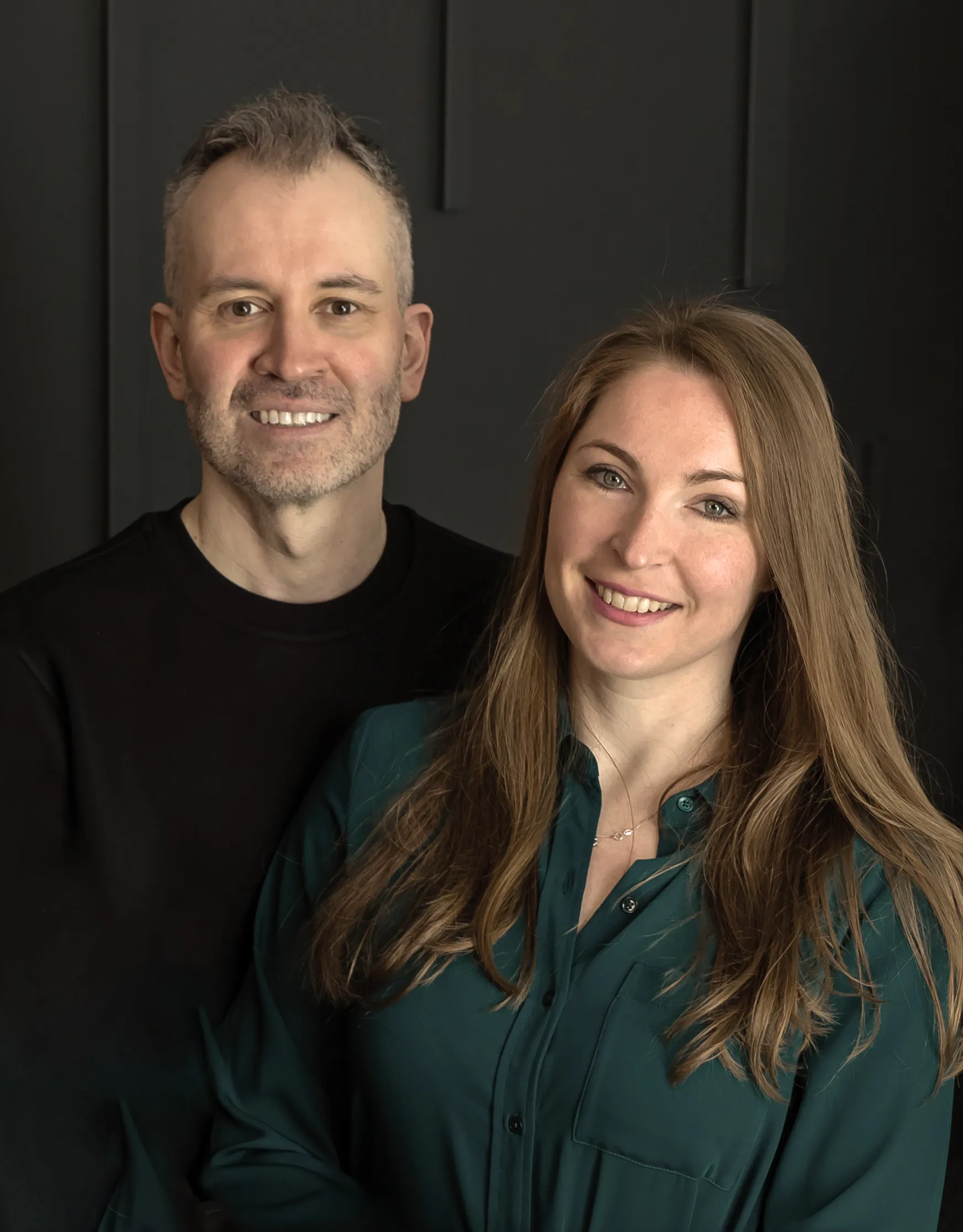 A sepia-toned professional portrait of a man and woman standing side by side, both smiling warmly at the camera. The man is on the left wearing a dark crew-neck sweater, with short cropped hair and light stubble. The woman is on the right with long wavy brown hair, wearing a dark button-up blouse and a delicate necklace. The background is a dark paneled wall, giving the photo a polished, studio-quality feel.